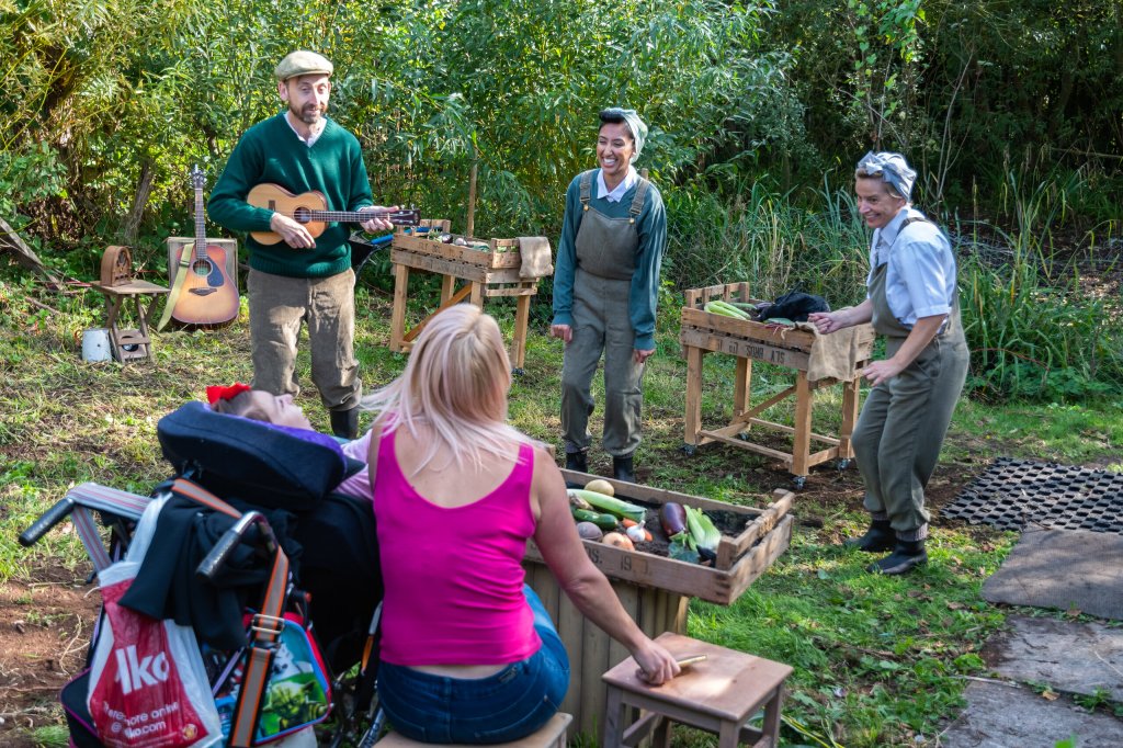 A photo taken of Bamboozle's Down to Earth show. A woman and a little girl in an electric wheelchair sit outside in, in front of a wooden box full of vegetables as 2 land girls and the allotment owner perform. The land girls also have a tray of vegetables and the allotment owner is playing a guitar.