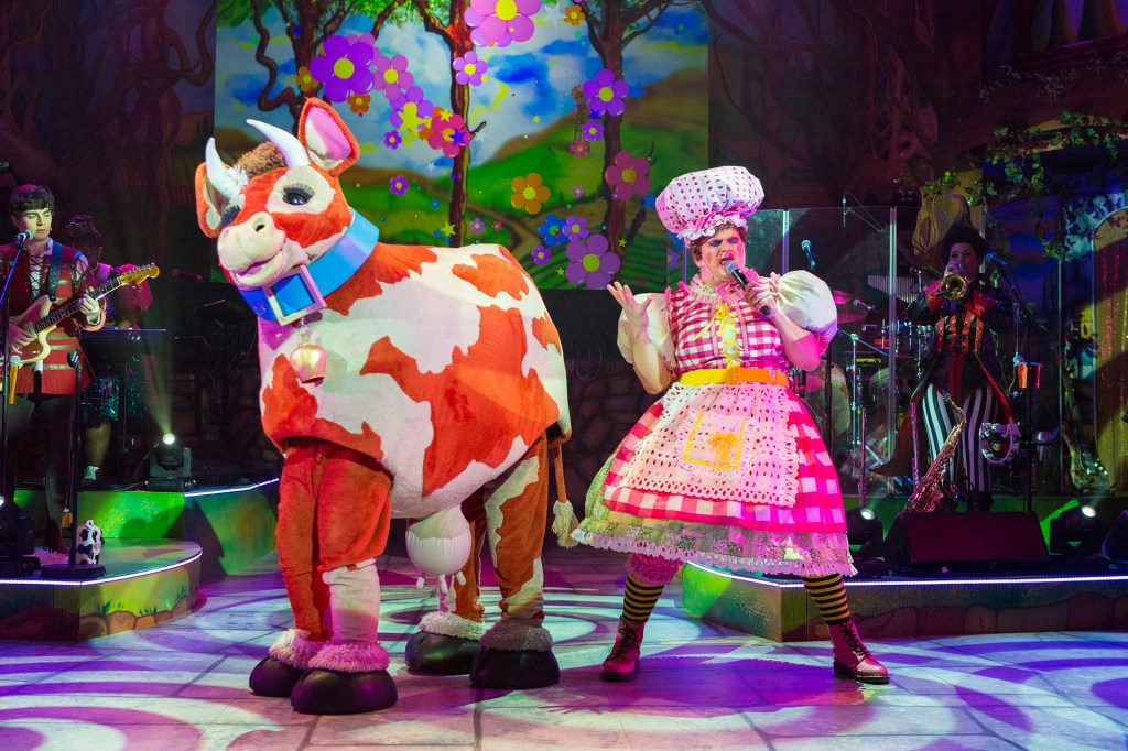 A production photo of Steve Simmonds as Dame Dolly Durden wearing a green patchwork dress under a pink and white gingham apron, green and black stripey tights, pink Doc Marten boots and a white with pink spots floppy hat singing with Bessie the cow. Behind them is a video screen showing flowers falling, Jack playing the guitar and Fleshcreep playing the trumpet. 